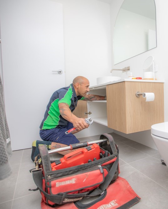 Plumber working underneath a bathroom sink | Featured image for the Maintenance Plumbers page of Akins Plumbing.